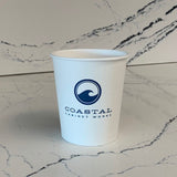 Custom printed 10 oz paper cup with "Coastal Cabinet Works" logo and wave circle graphic, on marble table
