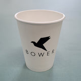 12 oz paper cup with black "Bower" logo and bird graphic, standing on light blue table