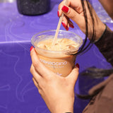 12 oz plastic cup filled with iced milk coffee, held by woman stirring with wooden stir stick over purple table