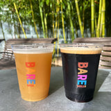 Two 16 oz plastic cups with "Barebottle Co" print, filled with light and dark beer with froth, on stone table, outside with benches and bamboo
