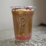 16 oz plastic cup with "Capo's Coffee" white logo, filled with milk coffee with rose additive and petals, standing on decorated table