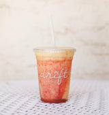 16 oz plastic cup with "Drift Palm Beach" white logo, filled with yellow drink and red syrup, with flat lid and white straw, on wooden table with white background