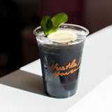 16 oz plastic cup with "Hustle Haven" orange logo, filled with dark drink and ice, garnished with lemon and lime, on white counter