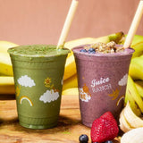 Two 16 oz plastic cups with "Juice Ranch" print, green and purple smoothies with paper straws, on wooden table with berries and bananas, pink background