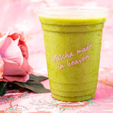 16 oz plastic cup with pink "Matcha Made in Heaven" logo, filled with green drink, on floral table with a rose to the left