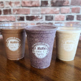 Three 16 oz plastic cups with white "Scenic City" logo and cityscape graphic, filled with smoothies, on wooden table, brick wall background