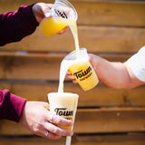 Three 16 oz plastic cups with "Town Brewery" black logo, filled with beer, with two people pouring one drink into another into the bottom cup