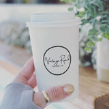 16 oz paper cup with white dome lid, printed "Vintage Road Coffee Co" circular logo, held by hand, table with plants in background