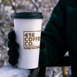 20 oz paper cup with blank dome lid and gold "416 Coffee Co" print, held by gloved hand, jacket sleeve visible, against metal railing, winter outdoors