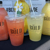 Two 20 oz plastic cups with flat lids and white straws, filled with pink and yellow lemonade, "Bold Quality Lemonade" logo printed, on black tablecloth with drink containers in background