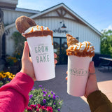 Two 20 oz paper cups with green "Grown and Bake" and "Pingle's" logos, caramel drinks with whipped cream, held by pink and green sweater hands, market entrance