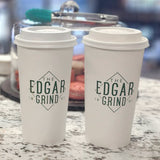 Two 20 oz paper cups with white dome lids and green "The Edgar Grind" print, on marble table, jar of macarons in background