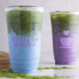 Two 24 oz plastic cups with pink "Spill the Tea" print and heart cup icon, filled with colorful drinks, on white table with matcha powder and whisk
