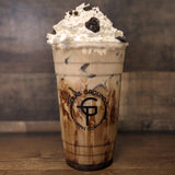 24 oz plastic cup with black "Texas Ground Coffee Company" text, filled with iced coffee, chocolate syrup, whip and cookie crumbs, on wooden table