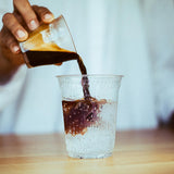 Hand pouring coffee into 7 oz printed plastic cup on table, with sparkling beverage