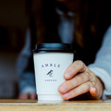 8 oz double-walled paper cup with "Amble Coffee" logo and bird print, black dome lid, held by hand with rings, on wooden table