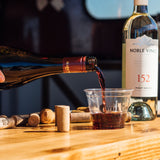9 oz custom printed plastic cup with Island Time logo, being poured with red wine, on wooden table with wine bottles and corks
