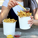 Two 24 oz paper food containers, filled with fries, one held by a woman, next to three portion cups filled with sauces on picnic table