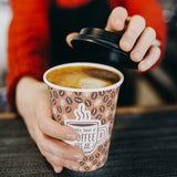 12 oz. Tan Full Wrap Recyclable Paper Cup, held on top of a wooden table, with the other hand taking off black lid