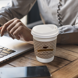12 oz paper cup with white dome lid and brown sleeve printed "Coffee Shop" graphic with cup icon, on table next to phone and person on laptop