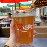 16 oz plastic cup with "LovePC" white logo, filled with dark orange beer, held by hand, outside patio bar with people in background