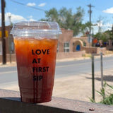20 oz plastic cup with "Love at First Sip" text, filled with orange iced tea, standing on metal railing with street in background
