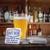 White coasters featuring the "Craft Beer logo", with one coaster standing upright against a beer glass on a wooden table at a bar.