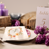 White paper sandwich bags with floral border and "KA" initials on a plate with a donut, decorated with purple flowers, wood, and a Table Five sign.