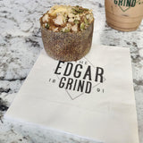 3-ply white beverage napkin featuring "The Edgar Grind" logo in black, placed on a greyscale marble table with a muffin resting on the napkin and an iced coffee in a takeout cup beside it.
