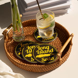 Four black round coasters, with yellow logo "Long Island Soda", in a woven tray, with a glass of lemonade and vase, on white table