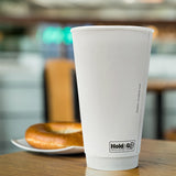 A 20 oz. white recyclable double walled paper cup is sitting on the surface of a table with a bagel in the distance. Cups are provided by The Cup Store Canada.