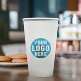 20 oz paper cup with blue "Your Logo Here" print, on wooden table, food assortment on shelving and plate with cookies in background