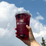 20 oz plastic cup with white "The Cupstore" print and sip lid, filled with black currant juice, berries and ice, held by hand with blue sky