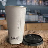 A 20 oz. white double walled paper cup and a black dome lid are next to each other. In the distance, is a blurred coffee shop menu and counter. Cups and lids are provided by The Cup Store Canada.