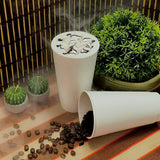 A steaming cup of coffee with whipped cream and chocolate drizzle sits upright while another lies on its side, spilling coffee beans. The scene is set on a table with a striped mat, featuring premium beverages in double-walled paper hot cups, alongside a miniature potted plant and some small cactus. Cups are provided by The Cup Store.