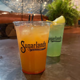Two 10 oz plastic cups with Sugarlands Distilling Co logo, filled with orange and green iced drinks with straws, on table