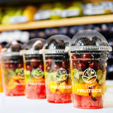 Four 10 oz plastic cups with The Fruitbox logo, filled with fruit and berries, on white table in grocery store