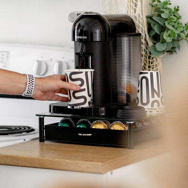 A person is using a Nespresso coffee machine, placing a 12 oz. custom-printed paper cup with a bold, modern design under the dispenser. A storage drawer below the machine holds various colored coffee pods. A watch is visible on the person's wrist. Nearby is a stove and a potted plant. The cups are printed by The Cup Store Canada.