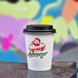 12 oz paper cup with black dome lid, green "Anthony & Son" text and red boy in car graphic, on metal table, colorful wall behind