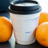 12 oz double-walled paper cup with black dome lid, blue pattern and "Clarity" text, standing on black table next to three oranges