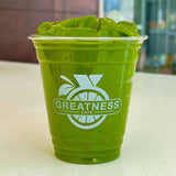 12 oz plastic cup filled with green smoothie, with "Greatness Cafe" and citrus logo, on white table with building in background