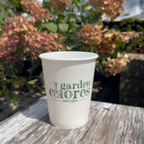 12 oz paper cup with green "Garden Chores and Coffee" logo, on wooden table, flowers and plants in background