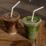 Two 12 oz plastic cups with Kettl logo, filled with brown and green drinks, flat lids, white straws, on wooden table with a cookie