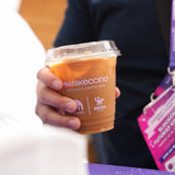 Milk coffee drink filled with ice in a cup with "Krakeccino" logo, held by person wearing black shirt and expo badge