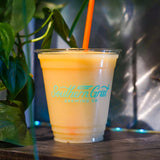 12 oz plastic cup filled with yellow slushy drink, with blue "Southern Grist" logo and orange straw, on wooden table with a plant