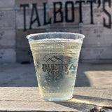 12 oz plastic cup with "Talbott's Cider" text and mountain logo, filled with light cider, on wooden table next to building with "Talbott's" sign