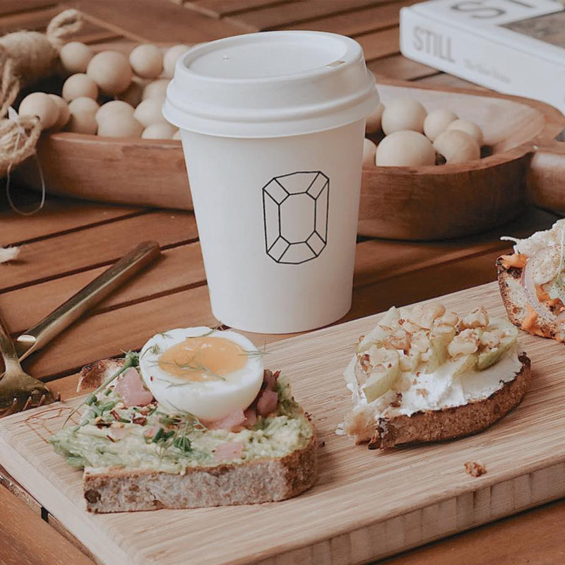 A 12 oz. custom-printed paper cup featuring a gem symbol stands on a wooden table next to an arrangement of open-faced sandwiches. The sandwiches include avocado toast with a soft-boiled egg and another topped with cream cheese and walnuts. A book and wooden beads lie in the background. The cups are printed by The Cup Store Canada.