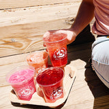 Four 16 oz plastic cups with "Familiars Coffee & Tea" white logo, filled with iced pink and red drinks, in cardboard carrier on wooden bench, person lifting one drink