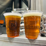 Two 16 oz plastic cups with "Boire Charlevoix" text and "Microbrasserie" logo, filled with frothy beer, standing on grey bar counter
