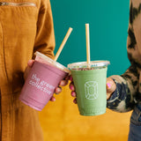 Two 16 oz plastic cups with flat lids and straws, left pink "The Green Collective" drink, right green smoothie with crystal logo, held by two women